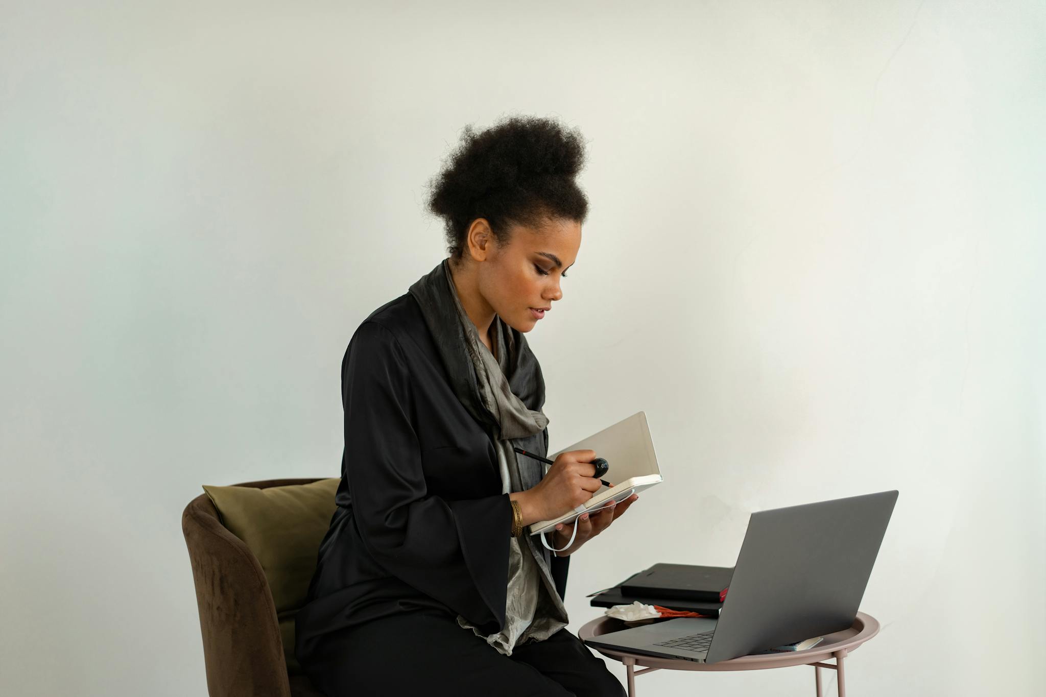 Woman in black dress taking notes while working remotely with a laptop indoors.